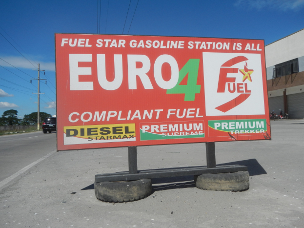 Tankstelle mit dem Schild "Euro4 Fuel", Fahrzeuge auf der Straße, Strommasten mit Drähten, Bäume, ein Gebäude auf der rechten Seite und sichtbarer Himmel.