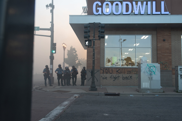 Eine Gruppe von Polizisten in Helmen mit Waffen vor einem Second-Hand-Laden mit Glaswänden und einer Tafel, mit Laternen und Verkehrszeichen im Vordergrund und Bäumen unter einem klaren Himmel im Hintergrund.
