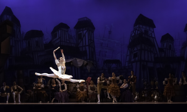 Eine Ballerina in einem weißen Tutu und Spitzenschuhen tanzt auf einer Bühne vor einem sitzenden und stehenden Publikum, mit einem Schlossgemälde im Hintergrund.