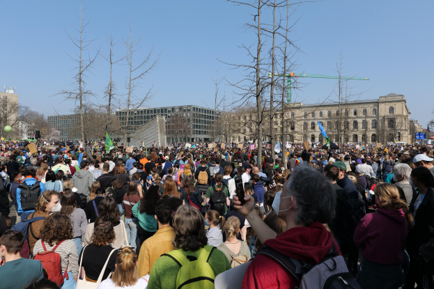 Eine große Menge von Menschen mit Schildern und Tötenschutzwesten vor einem Gebäude mit Fenstern, Bäumen und einem klaren Himmel im Hintergrund, was auf eine Klimawandeldemonstration hindeutet.