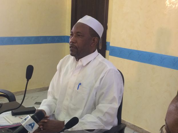 Ein Mann in einem weißen Gewand und Hut sitzt an einem Tisch mit Mikrofonen, spricht in einem Raum mit einer anderen Person zu seiner Rechten, Papieren auf dem Tisch und einer Tür im Hintergrund.