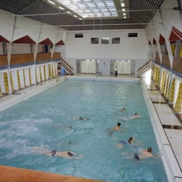 Großes Hallenbad mit schwimmenden Menschen, flankiert von Säulen, mit einer Wand mit Türen und einer Uhr im Hintergrund und Deckenlampen, die den Raum beleuchten.