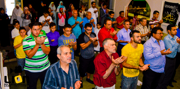 Eine Gruppe von Menschen betet in einem großen Saal, mit Text in der linken unteren Ecke.