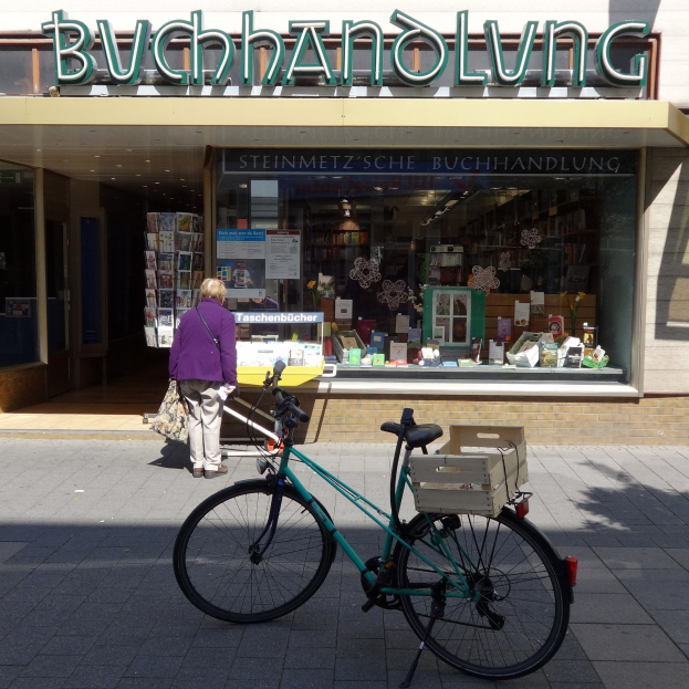 Eine Frau steht neben einem Fahrrad, das vor einem Laden geparkt ist, mit einem Fahrradrad im Vordergrund und durch die Glasscheibe des Ladens sichtbaren Büchern.