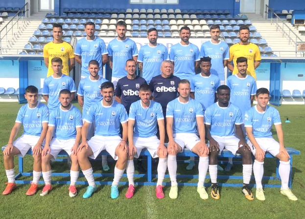 Eine Gruppe von Männern, einige stehen, einige sitzen auf Bänken, auf einem Fußballfeld, mit Stühlen, Treppen, Geländern, Wänden und Fenstern im Hintergrund.