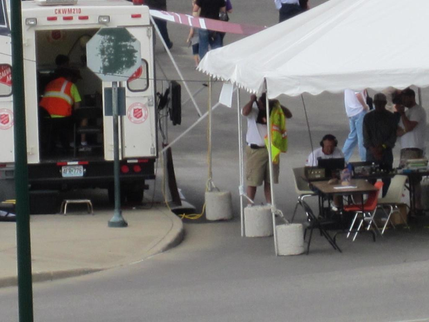 Ein Krankenwagen steht links von einer Straße, während auf der rechten Seite ein Zelt viele Menschen beherbergt, mit einem Tisch und einem Stuhl in der Nähe.