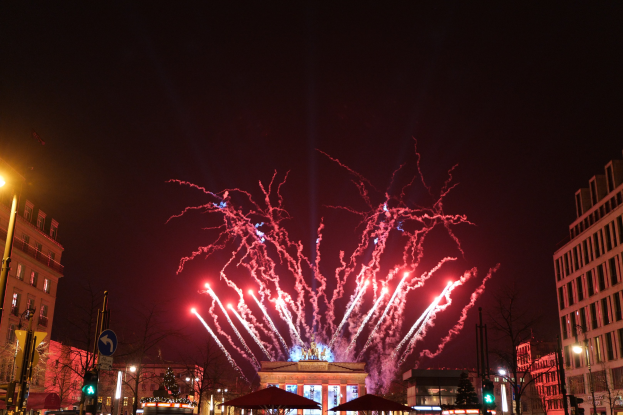 Eine belebte Stadtstraße in Berlin am Neujahrstag, voller Menschen, Fahrzeuge und festlicher Dekorationen, mit Feuerwerk, das den Nachthimmel erhellt.