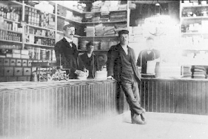 Ein Schwarz-Weiß-Foto von drei Männern, die an einem Tresen in einem Laden stehen, mit verschiedenen Gegenständen auf dem Tresen und einem Regal, das mit Büchern und anderen Gegenständen gefüllt ist, während eines Überfalls.