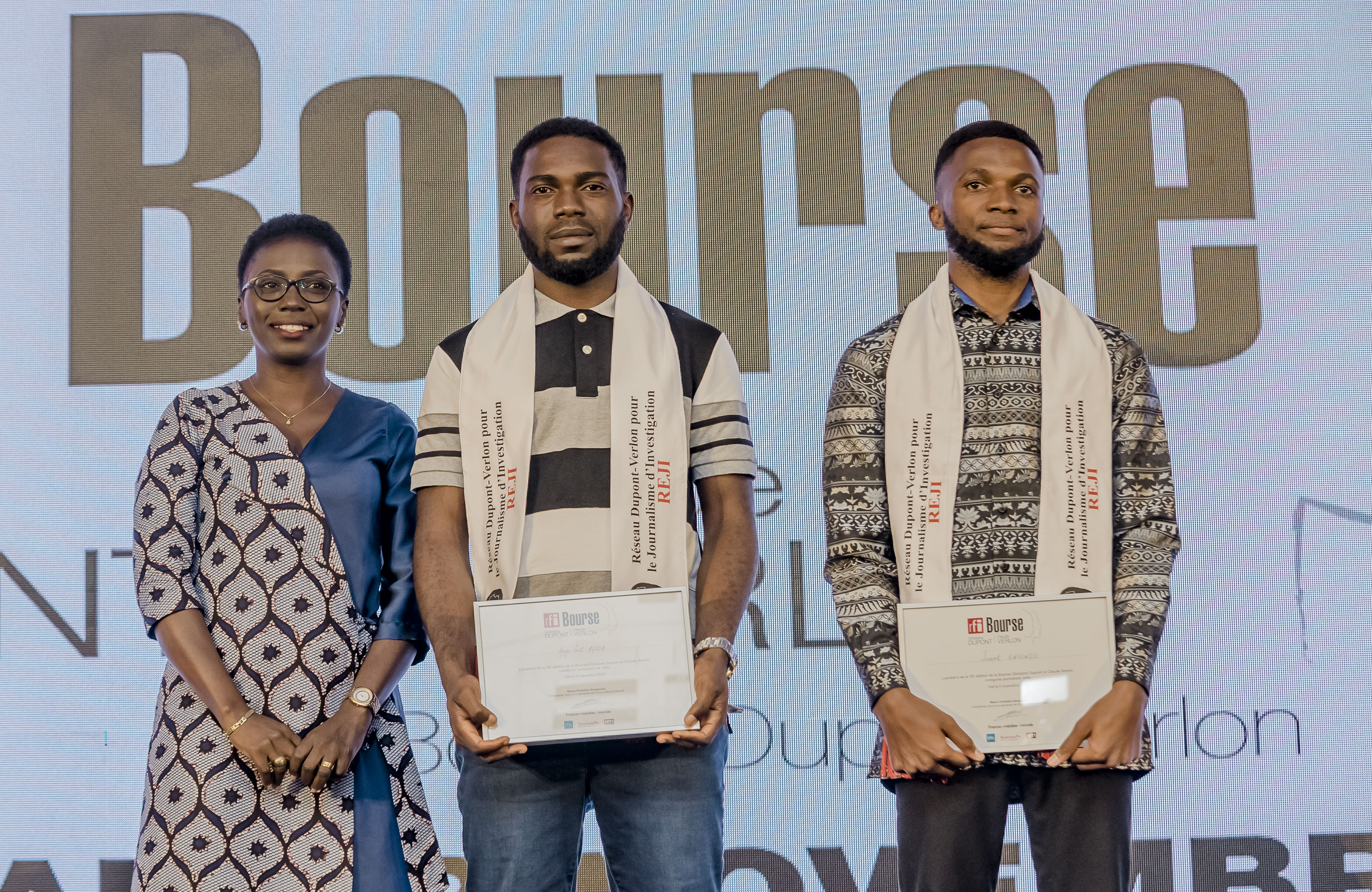 Drei Personen stehen nebeneinander auf einer Bühne und halten jeweils ein Zertifikat in der Hand, im Hintergrund ist ein Bildschirm mit der Aufschrift "Bounce Africa Awards 2019" zu sehen.