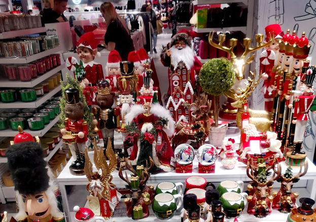Ladeninnenraum weihnachtlich dekoriert mit Tischen voller Spielzeug, Tassen und anderen Gegenständen, Regalen mit Waren und Menschen im Hintergrund bei beleuchteten Auslagen und einer Wand.