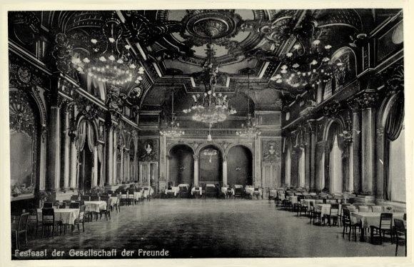 Ein altes Schwarz-Weiß-Foto eines großen Raums mit Tischen, Stühlen, Säulen, Bögen und Kronleuchtern an der Decke, mit Text unten, der "Fest der Gesellschaft der Freunde, Berlin" lautet.