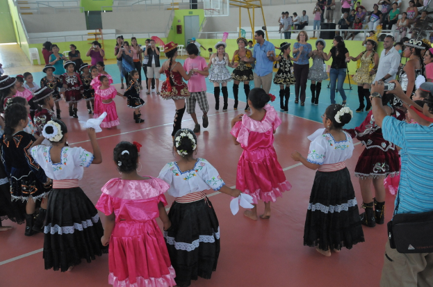 Eine Gruppe von Kindern in Kostümen tanzt in der Mitte des Bildes, während das Publikum von der oberen rechten Seite zusieht.