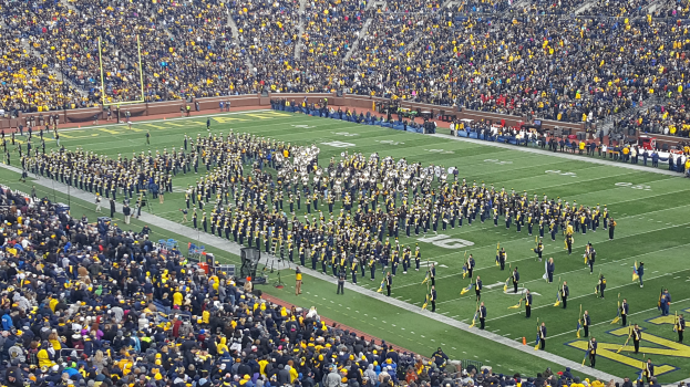 Eine große Menschenmenge in einem Stadion bei einem Fußballspiel, wobei einige stehen und Fahnen schwenken, andere sitzen und Pfosten und Geländer im Hintergrund zu sehen sind.