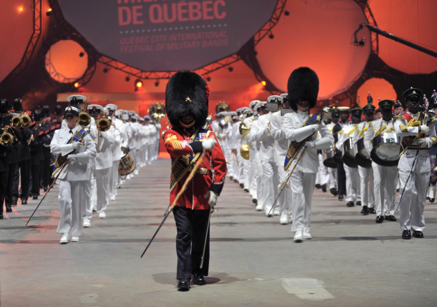 Eine Gruppe uniformierter Personen marschiert durch die Straßen, einige spielen Musikinstrumente, mit einem beleuchteten Schild im Hintergrund, das die Eröffnungszeremonie des Montreal International Festival of Military Bands anzeigt.