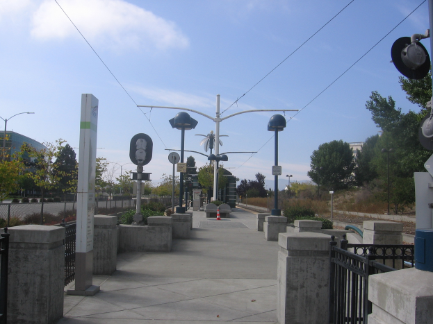 Ein Fußgängerbrücke mit Geländern, Pfählen und Laternen, umgeben von Pflanzen, Bäumen und Gebäuden, mit einem Zug auf den Gleisen in der Nähe unter einem bewölkten Himmel.
