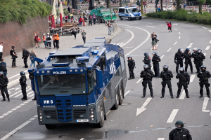 Gruppe von Polizisten in Uniform vor einem gepanzerten Fahrzeug auf einer Straße mit bewaffneten Personen in Helmen und Zivilisten im Hintergrund bei Gebäuden und Verkehrszeichen.