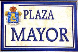 Ein rechteckiges weißes Schild mit schwarzer Schrift, die "Plaza Mayor" lautet, und einem blauen Rahmen, das ein gekröntes Wappen-Logo trägt und an einem Gebäude angebracht ist.