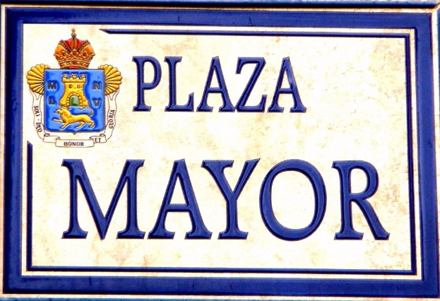 Ein rechteckiges weißes Schild mit schwarzer Schrift, die "Plaza Mayor" lautet, und einem blauen Rahmen, das ein gekröntes Wappen-Logo trägt und an einem Gebäude angebracht ist.