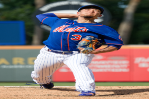 Ein Baseballspieler in einem blauen T-Shirt, weißen Hosen, einer blauen Mütze und blauen Schuhen wirft einen Ball auf einem Feld mit einer Werbetafel und Bäumen im Hintergrund.