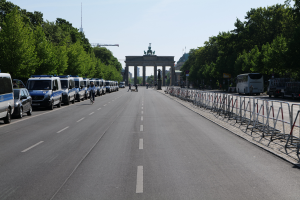 Eine lange Reihe von Polizeiwagen, die auf der Seite einer Straße vor dem Brandenburger Tor in Berlin, Deutschland, geparkt sind, mit Menschen, die Fahrräder fahren und auf der Straße stehen, Absperrungen und Bäume, die die Seiten säumen, und ein Tor mit Statuen im Hintergrund.