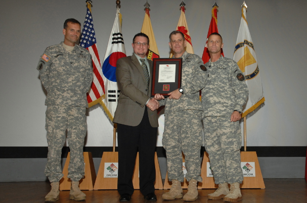 Vier Männer stehen auf einer Bühne: drei in Armeeuniformen und einer in Blazer, Krawatte und Hemd, die Hände schüttelnd und einen Preis überreichend, mit sechs Fahnen auf Stangen und einer weißen Wand im Hintergrund.