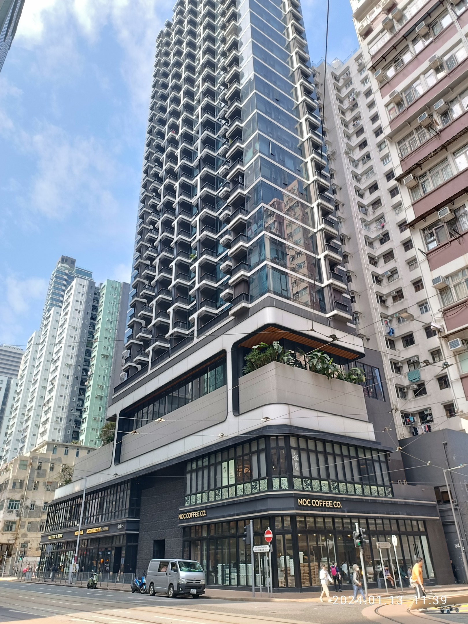 Ein hohes Gebäude mit der Aufschrift "Noc Coffee Co." auf einer belebten Stadtstraße umgeben von anderen Gebäuden, Straßeninfrastruktur, Fahrzeugen, Fußgängern, Bäumen und einem bewölkten Himmel.