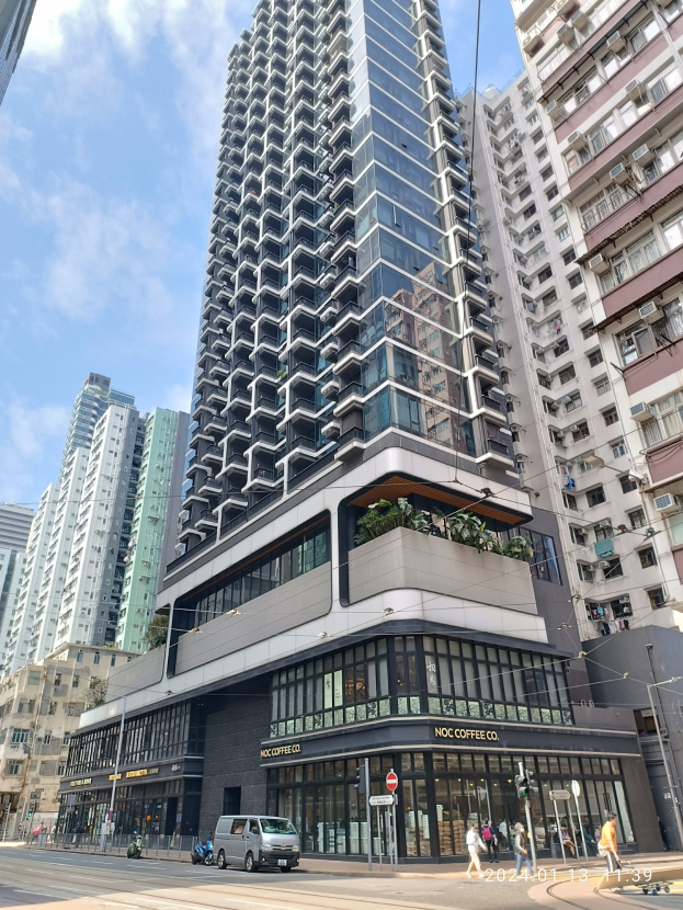 Ein hohes Gebäude mit der Aufschrift "Noc Coffee Co." auf einer belebten Stadtstraße umgeben von anderen Gebäuden, Straßeninfrastruktur, Fahrzeugen, Fußgängern, Bäumen und einem bewölkten Himmel.