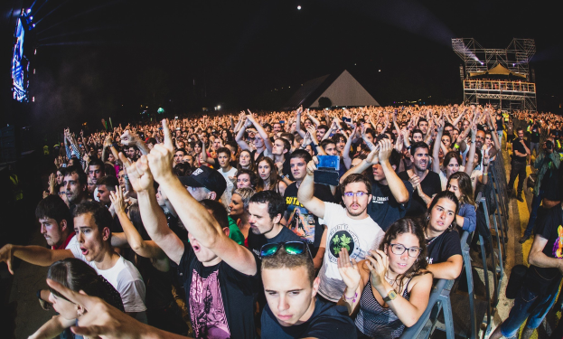 Menschenmenge auf einem Musikfestival mit erhobenen Händen, einige sitzen und andere stehen, von einem Metallzaun umgeben, mit Gebäuden und Lichtern im Hintergrund unter einem dunklen Himmel.
