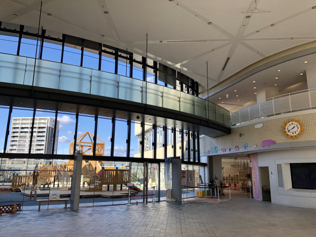 Innenansicht eines großen Gebäudes mit hohen Decken, zahlreichen Fenstern, elektrischem Licht, einer Wanduhr, einem Fernseher, einem Bildschirm, hölzernen Strukturen, einem hölzernen Zaun, benachbarten Gebäuden, Bäumen und einem bewölkten Himmel, das als Haupteingang zu einem neuen Kinderspielbereich dient.