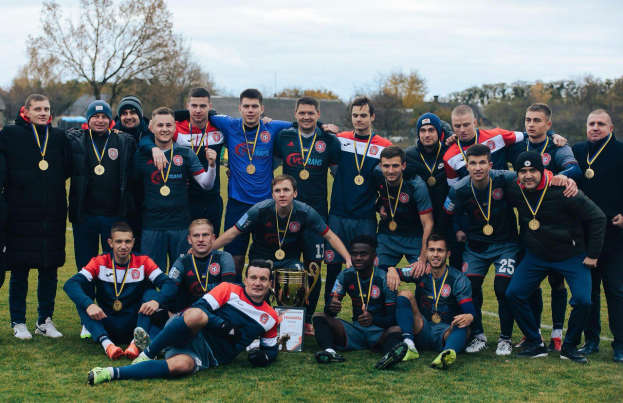 Eine Gruppe von Männern in Medaillen steht auf einem Fussballfeld und hält einen Pokal, umgeben von Bäumen unter einem klaren blauen Himmel.