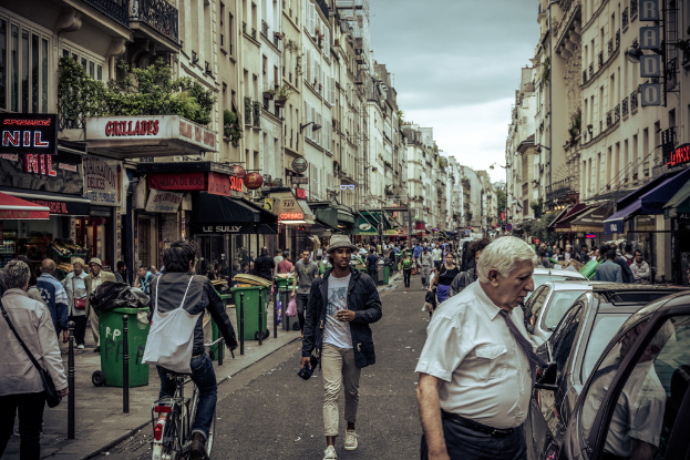 Zwei große Gebäude mit Geschäften und Mülltonnen im Erdgeschoss, getrennt durch eine belebte Straße mit Fußgängern, einem Radfahrer und parkenden Autos.
