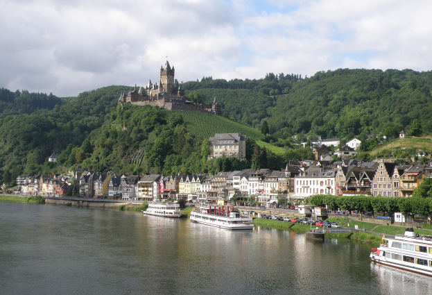 Ein malerischer Blick auf den Rhein in Deutschland mit einer Burg auf einem Hügel, Booten auf dem Fluss und Fahrzeugen auf der Straße sowie einem bewölkten Himmel.