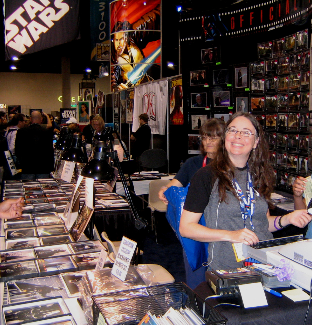 Eine Frau posiert an einem Zahlungstresen in einem belebten Geschäft, das mit Bildern, Büchern und ausgestellten Plakaten gefüllt ist, während Kunden im Hintergrund einkaufen.