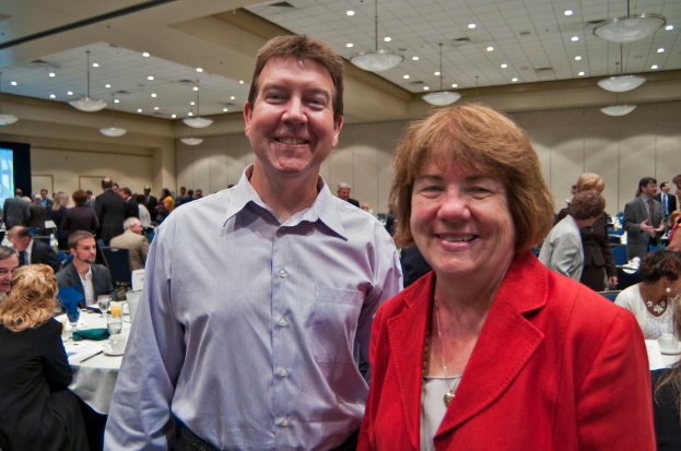 Zwei Personen stehen lachend im Vordergrund, während andere an Tischen mit Tassen und Gläsern sitzen und weitere Personen im Hintergrund an einer Wand diskutieren.