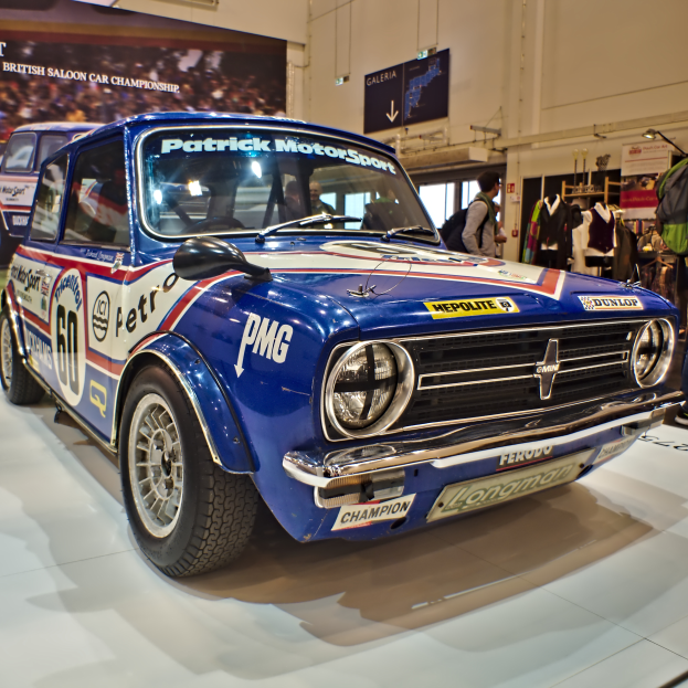 Ein kleiner blauer und weißer Mini Cooper S Rallye-Wagen, der in einem Museum ausgestellt ist und von Menschen umgeben ist, die Taschen tragen und um ihn herumstehen. Im Hintergrund befindet sich eine Wand mit einem Banner, das Text und Bilder zeigt, sowie Lichter und andere Gegenstände.