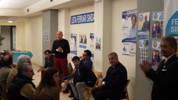 Eine Gruppe von Menschen, mit zwei stehenden und dem Rest sitzend, vor einer Wand mit verschiedenen Textplakaten.