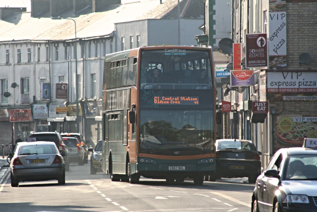 Eine Straße mit Autos und einem Bus, Gebäude mit Wänden, Fenstern, Tellern und Dächern, Plakate und Banner an den Wänden und ein Pfahl mit einer Straßleuchte.