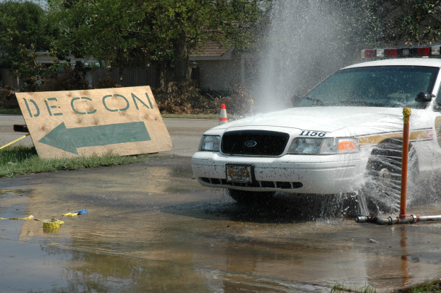 Ein Streifenwagen fährt durch eine Pfütze, mit einem Schild, einem Verkehrskegel und anderen Gegenständen auf dem Boden, vor einem Hintergrund aus Bäumen, Gebäuden und einem klaren blauen Himmel.