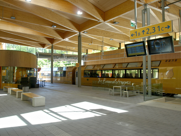 Ein belebter Bahnhof mit einem Zug auf den Gleisen, umgeben von Bänken, Tischen, Pfählen, Schildern, Laternen und Bäumen und voller Menschen, die auf den Zug warten.