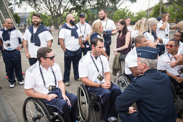 Eine Gruppe von Menschen im Rollstuhl vor einem Hintergrund aus Bäumen, Pfählen, Gebäuden und einem klaren blauen Himmel, wobei einige Mützen und Brillen tragen.