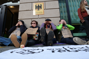 Eine Gruppe von Menschen sitzt auf dem Boden und hält Schilder, mit einer Fahne und einem Apfel auf der Straße, vor einem Gebäude mit Glasfenstern und einer Tür, das besorgt aussieht.