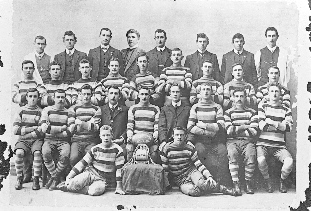 Schwarzes und weißes Foto einer Fußballmannschaft mit sitzenden und stehenden Spielern, einem Helm in der Mitte und einem leicht unscharfen Hintergrund.