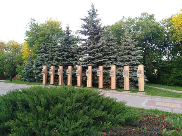 Ein Park mit einer Reihe von Bäumen in der Mitte, umgeben von blühenden Pflanzen, Gras, einem Weg, einem Straßenmast und einer Gruppe von Bäumen unter einem bewölkten Himmel, mit einem Gedenkschild für die Opfer von Tschernobyl.