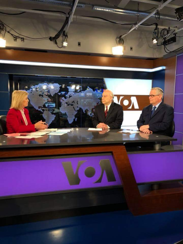 Drei Personen, zwei Männer in Blazern und Krawatten und eine Frau in einem roten Kleid sitzen an einem Schreibtisch mit Papieren und Gegenständen, die in eine Diskussion vor einem Fernseher vertieft sind, mit einem Bildschirm und Lampen im Hintergrund.