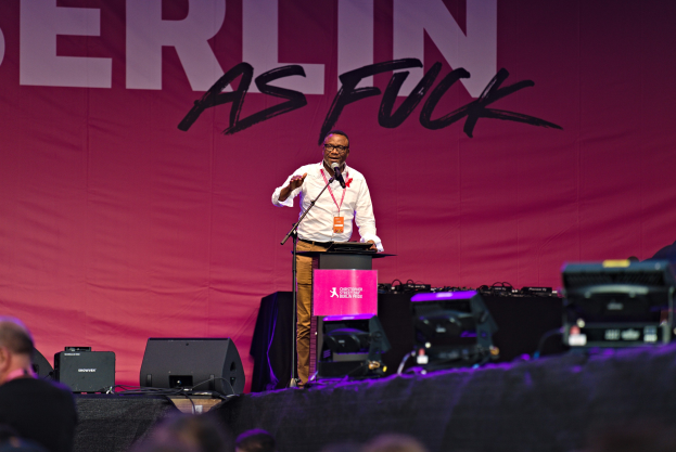 Ein Mann in einem weißen Hemd und braunen Hosen steht auf einer Bühne und hält ein Mikrofon in der Hand, während er vor einem pinken Banner mit der Aufschrift 'Berlin as Fuck' spricht.