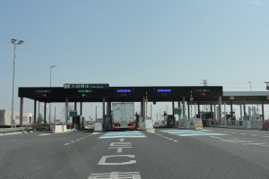 Mautstelle mit einem vorfahrenden Lastwagen auf einer stra├čen mit wei├čen und blauen Markierungen, Leuchtmasten, Schildern und anderen Gegenst├Ąnden, mit Geb├Ąuden, B├Ąumen und einem klaren blauen Himmel im Hintergrund.