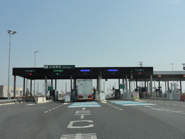 Mautstelle mit einem vorfahrenden Lastwagen auf einer stra├čen mit wei├čen und blauen Markierungen, Leuchtmasten, Schildern und anderen Gegenst├Ąnden, mit Geb├Ąuden, B├Ąumen und einem klaren blauen Himmel im Hintergrund.