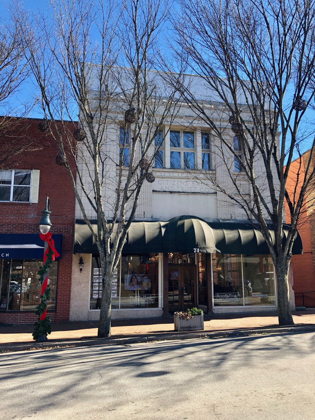 Ein Eckgebäude mit einer grünen Markise, Glasfenstern und einer Namenstafel, auf der 'Citizens Bank and Trust Co.' steht, ist von Bäumen und einem Laternenpfahl umgeben und hat einen bewölkten Himmel im Hintergrund.