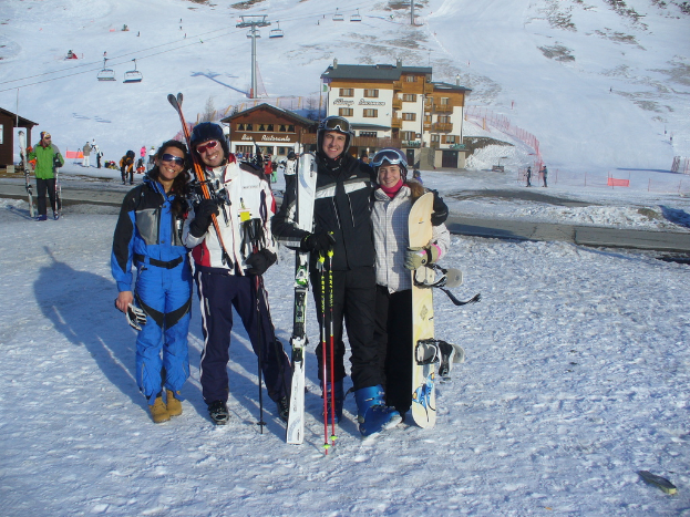 Vier Personen posieren mit Schlittschuhausrüstung und Werkzeugen für ein Foto, mit Gebäuden und anderen Menschen im Hintergrund, vor einer schneebedeckten und vereisten Berglandschaft.