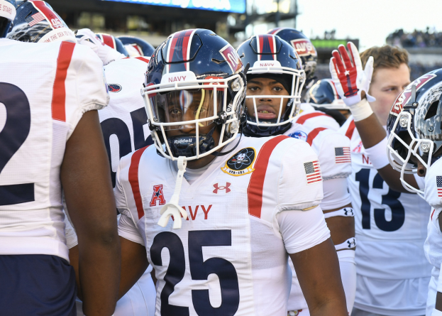 Eine Gruppe von Männern in Football-Uniformen steht auf einem Feld mit einer unscharfen Menge, einem Scoreboard mit 'Auburn vs Virginia Tech - 11/28/15 College Football Pick, Odds, and Prediction' und Himmel im Hintergrund.